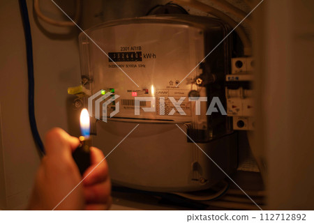 A man's hand with a lighter in complete darkness looks at the electric meter at home. Power outage, blackout concept. 112712892
