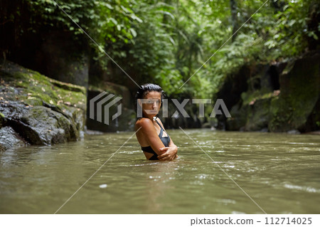 A young slender woman in a swimsuit poses by a mountain river in the jungle on the popular island of Bali. 112714025