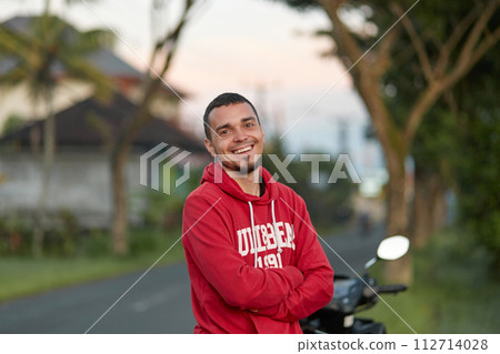Portrait of a young man with a beard, smiling. A guy in a red sweatshirt is standing against the sunset. Portrait of a young man with a beard, smiling. A guy in a red sweatshirt is standing against the sunset. 112714028