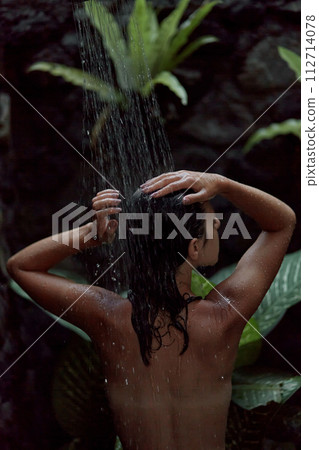 A naked woman takes a shower in an outdoor shower with tropical plants. A shampoo advertisement in dark colors. 112714078