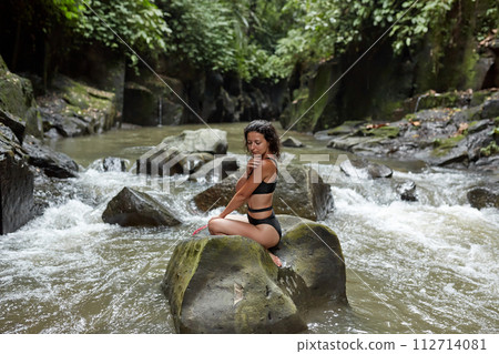 A young slender woman in a swimsuit poses on a huge boulder covered with moss in the middle of a mountain river in the jungle on the popular island of Bali. 112714081
