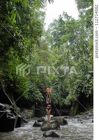 A young slender woman in a swimsuit poses with a large dry leaf on a huge boulder covered with moss in the middle of a mountain river in the jungle on the popular island of Bali. A young slender woman in a swimsuit poses with a large dry leaf on a huge boulder covered with moss in the middle of a mountain river in the jungle on the popular island of Bali. 112714082