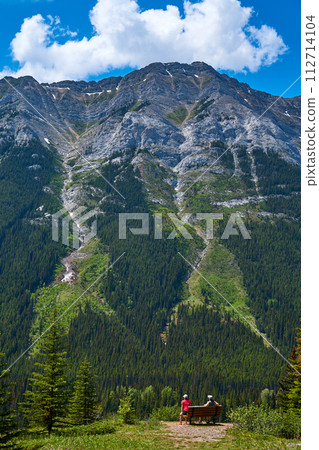 A couple sits on a bench and enjoys the view of a majestic rocky mountain in the Alberta province of Canada. 112714104