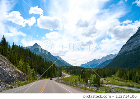 Beautiful view from a car on the Rocky Mountains in Banff National Park in Alberta. Panorama of a road in the mountains past a coniferous forest on a sunny day in summer. 112714106