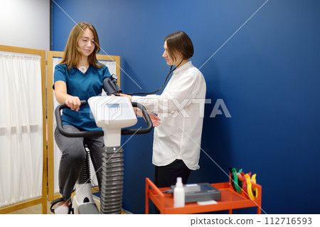Female doctor cardiologist making bicycle electrocardiogram for examination cardiovascular system of patient at medical office of clinic. Young woman training on bike simulator. 112716593