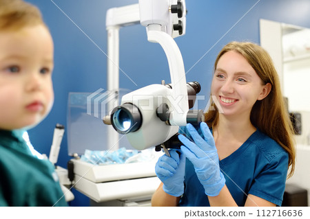 A cute toddler boy with his mother are at an appointment with a pediatric ENT to prepare for preschool. The doctor otolaryngologist is examining the ear of a little patient under ENT microscope A cute toddler boy with his mother are at an appointment with a pediatric ENT to prepare for preschool. The doctor otolaryngologist is examining the ear of a little patient under ENT microscope 112716636