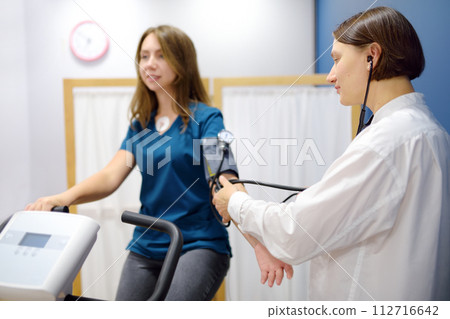 Female doctor cardiologist measures pressure with patient during bicycle exercise for examination cardiovascular system at medical office of clinic. Young woman training on bike simulator. 112716642