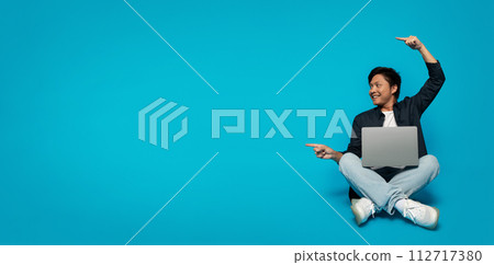 Smiling young man sitting cross-legged with a laptop, pointing and looking upwards towards copy space Smiling young man sitting cross-legged with a laptop, pointing and looking upwards towards copy space 112717380