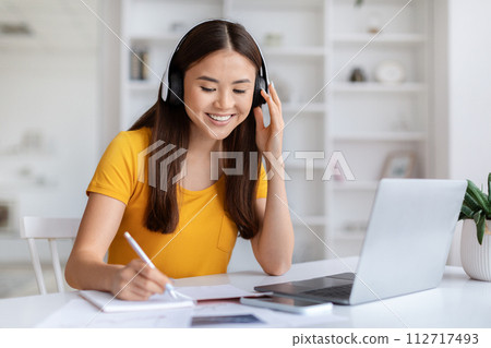 Smiling, young asian woman wearing headphones taking notes and using her laptop 112717493