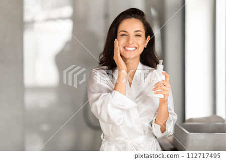 Cheerful young brunette woman holding bottle with face cream Cheerful young brunette woman holding bottle with face cream 112717495