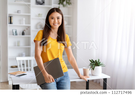 Smiling young asian woman holding closed laptop, standing in well-lit home office 112717496