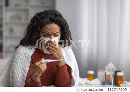 Sick Black Woman Blowing Runny Nose And Looking At Thermometer At Home Sick Black Woman Blowing Runny Nose And Looking At Thermometer At Home 112717695