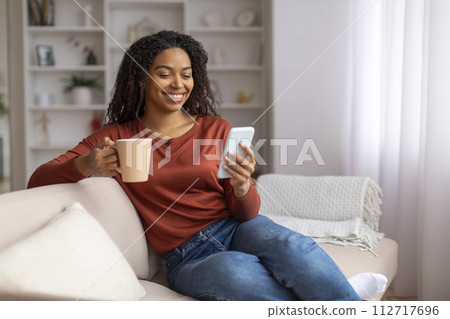 Young black woman relaxing on couch with coffee mug and smartphone 112717696