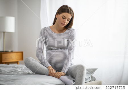 Young pregnant woman in casual wear massaging her swollen ankle at home 112717715