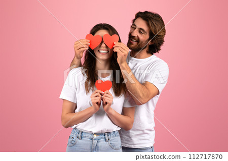 Joyful couple playfully covers their eyes with red paper hearts, sharing a light-hearted moment together Joyful couple playfully covers their eyes with red paper hearts, sharing a light-hearted moment together 112717870