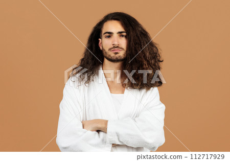 Confident Hispanic young man in bathrobe crosses arms, beige backdrop 112717929