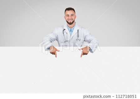 Doctor man pointing at empty white board below, grey background Doctor man pointing at empty white board below, grey background 112718051