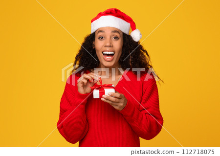 Surprised black lady with Santa hat, holds small gift, yellow background Surprised black lady with Santa hat, holds small gift, yellow background 112718075