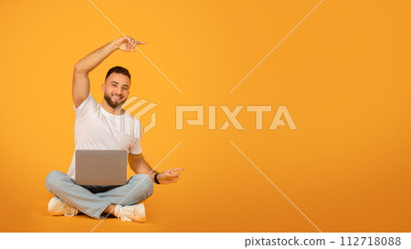 Smiling man in a white t-shirt and light blue jeans sitting cross-legged on the floor with a laptop 112718088