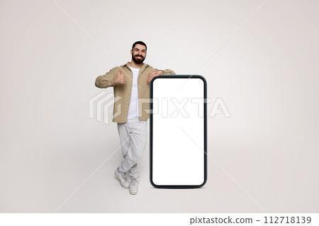 A cheerful man with a beard and a casual beige shirt stands giving a thumbs up beside a gigantic smartphone 112718139