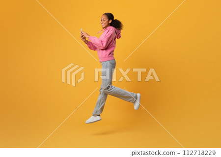 black teen girl leaping in air using phone, yellow background 112718229