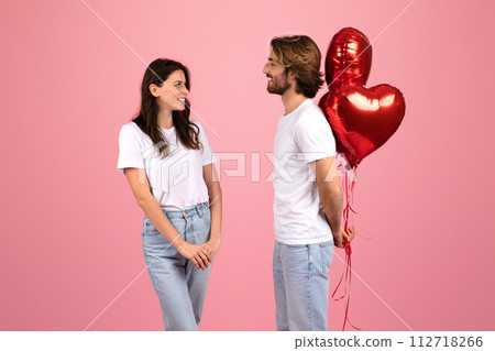 A happy couple stands facing each other with a playful expression, holding shiny red heart-shaped balloons A happy couple stands facing each other with a playful expression, holding shiny red heart-shaped balloons 112718266