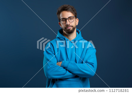 Man wearing glasses and blue hoodie looking disinterested, standing with arms crossed Man wearing glasses and blue hoodie looking disinterested, standing with arms crossed 112718281