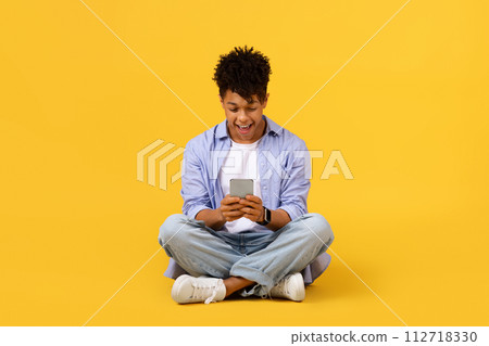 Joyful black guy using smartphone while sitting cross-legged on yellow background 112718330