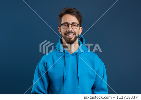 Cheerful young man with beard and glasses standing against dark blue background 112718337