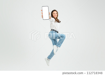 Dynamic young woman in mid-air jump, joyfully presenting a smartphone with a blank screen Dynamic young woman in mid-air jump, joyfully presenting a smartphone with a blank screen 112718348