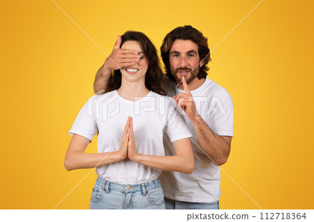 Playful man with a beard covering a woman's eyes with his hand, making a shushing gesture with finger on lips Playful man with a beard covering a woman's eyes with his hand, making a shushing gesture with finger on lips 112718364