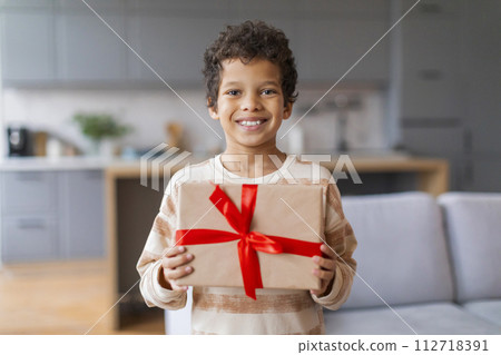 Happy little black boy proudly holding wrapped gift with bright red ribbon Happy little black boy proudly holding wrapped gift with bright red ribbon 112718391