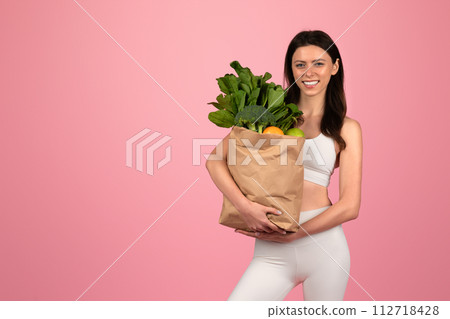Healthy lifestyle concept with a cheerful young woman in white athletic wear holding a grocery bag 112718428