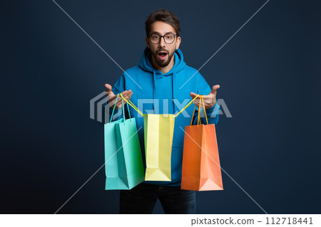 Shocked young man wearing glasses holding multiple colorful shopping bags Shocked young man wearing glasses holding multiple colorful shopping bags 112718441