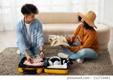 Engaged African American couple sitting on the floor, joyfully packing clothing into a bright yellow suitcase 112718461