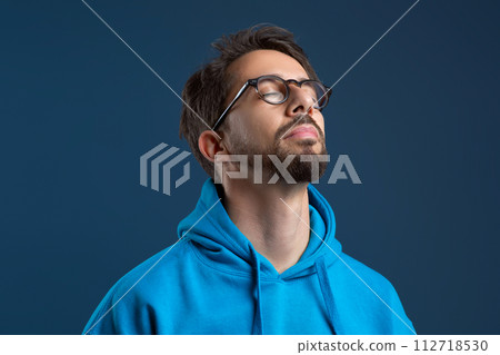 Serene young man wearing glasses closing his eyes in peaceful pose Serene young man wearing glasses closing his eyes in peaceful pose 112718530