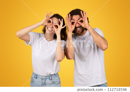 Playful couple making funny glasses with fingers over eyes, laughing in matching white t-shirts Playful couple making funny glasses with fingers over eyes, laughing in matching white t-shirts 112718596