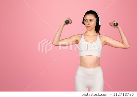 Focused young woman in white athletic wear flexing muscles with dumbbells Focused young woman in white athletic wear flexing muscles with dumbbells 112718756
