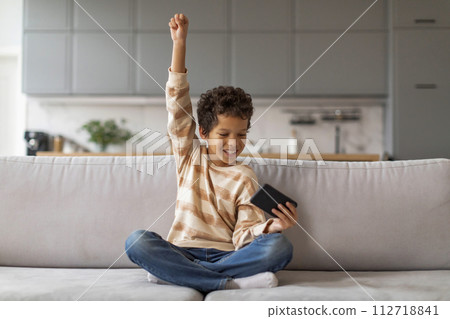 Joyful black boy holding smartphone and raising his arm in victory 112718841