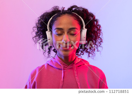 Content black lady enjoying music with in headphones, pink hoodie, and closed eyes Content black lady enjoying music with in headphones, pink hoodie, and closed eyes 112718851
