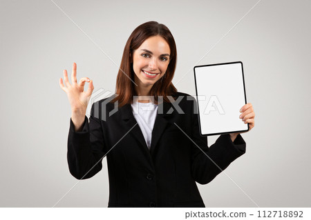 A poised young businesswoman holds a tablet with a blank screen in one hand while making an 'okay' A poised young businesswoman holds a tablet with a blank screen in one hand while making an 'okay' 112718892