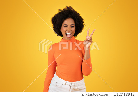 Vivacious young woman with fluffy afro hair winks and flashes a peace sign 112718912