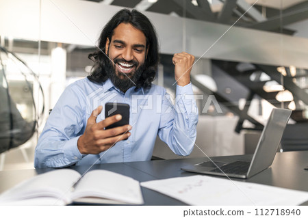 Joyful Young Indian Businessman Celebrating Success on Smartphone 112718940