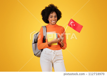 Joyful young woman with curly hair holding a Turkish flag, with headphones and a backpack 112718970