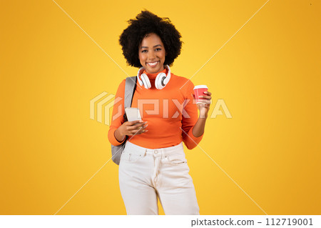 Smiling young woman multitasking with headphones, holding a coffee cup Smiling young woman multitasking with headphones, holding a coffee cup 112719001