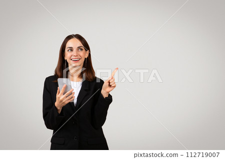 Joyful young businesswoman holding a smartphone and pointing upwards 112719007