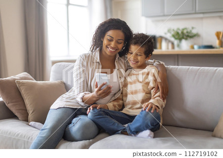 Family gadgets. Happy black mother showing new application on smartphone to son Family gadgets. Happy black mother showing new application on smartphone to son 112719102