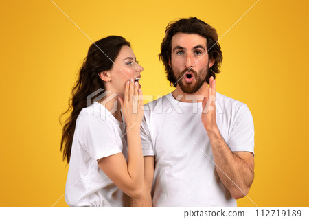 Surprised bearded man with wide eyes and an open mouth holds his hands up while a woman whispers into his ear Surprised bearded man with wide eyes and an open mouth holds his hands up while a woman whispers into his ear 112719189
