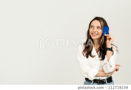 Thoughtful young woman holding a blue credit card to her temple, in a white blouse Thoughtful young woman holding a blue credit card to her temple, in a white blouse 112719239
