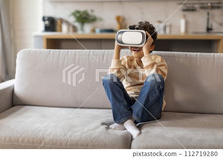 Excited black boy wearing virtual reality headset sitting on couch at home 112719280